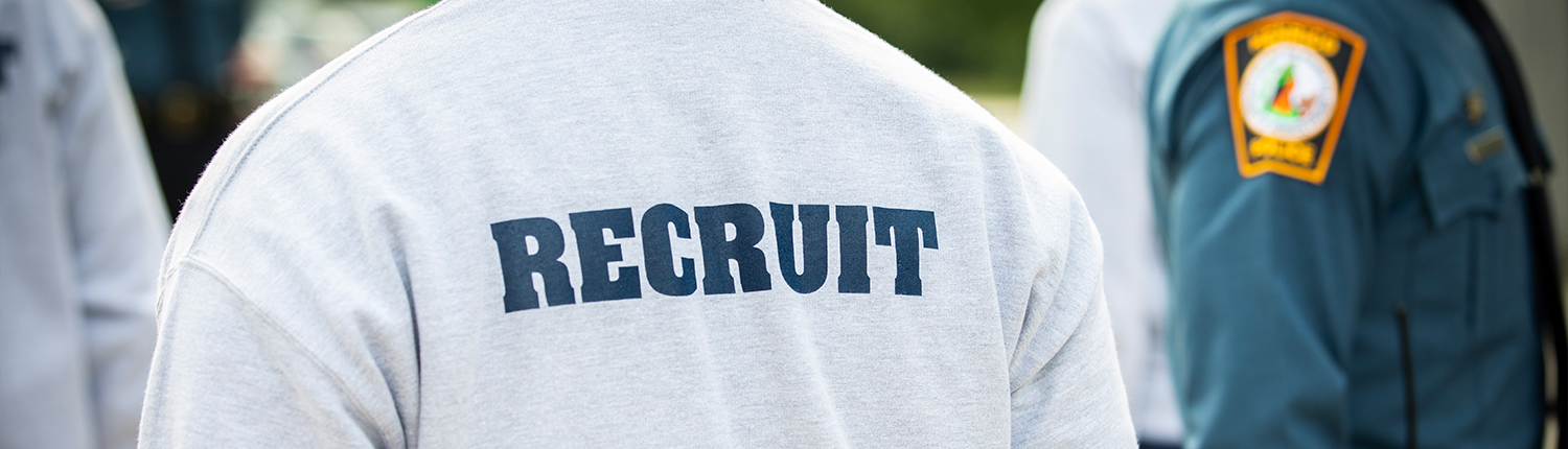 The back of a police recruit's shirt and a police in the background walking by.